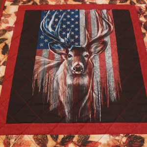 May include: A red, white, and blue American flag with a white-tailed deer in front of it. The deer has large antlers and is looking directly at the viewer. The flag is faded and distressed, giving it a vintage look.