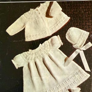 May include: Cream-colored baby clothing set, including a jacket, dress, bonnet, and booties. The set features intricate knitted detailing and a scalloped edge. The image includes the text "COPLEY LOTUS" and "Baby Golden Sovereign 4 ply". The size is 18 inches (46 cm).