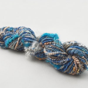May include: A skein of hand-spun yarn in shades of blue, white, and beige. The yarn is made with a variety of fibers, including wool, silk, and mohair. It has a unique, textured look and feel.