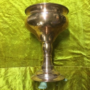 May include: A large, antique, brass planter with a wide, round top and a tall, slender base. The planter has three decorative feet.