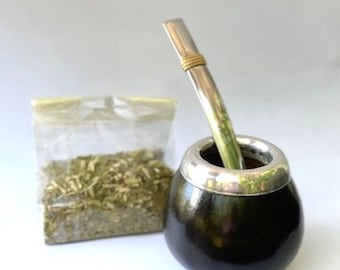 May include: A dark brown, rounded mate gourd with a polished silver rim, accompanied by a silver bombilla straw featuring a gold-colored band. Beside the gourd sits a clear plastic bag filled with dried, loose-leaf green yerba mate tea.