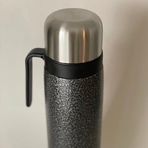 Puede incluir: Un termo de acero inoxidable con asa negra y acabado gris texturizado.