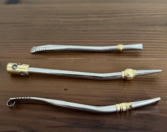 May include: Three silver-toned metal bombilla straws for drinking mate, each featuring a slightly curved stem and distinct gold-toned decorative accents. The straws vary in design, with one showing a perforated filter end and another a looped tip.