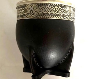 May include: A black mate gourd cup featuring a silver-colored metal rim with an embossed pattern of fleur-de-lis symbols and intricate braided detailing. The cup is supported by a black leather base with four distinct, stitched structural legs.