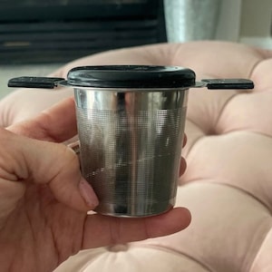 May include: A stainless steel tea infuser with a black lid and two handles. The infuser has a fine mesh design.