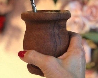May include: A brown ceramic mate gourd with a metal bombilla straw. The gourd is being held by a hand with red nail polish.