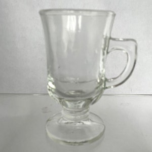 May include: Clear glass Irish coffee mug with a handle and a pedestal base. The mug has a flared top and a rounded handle. The glass is transparent, allowing the contents to be visible. The mug is designed for hot beverages.