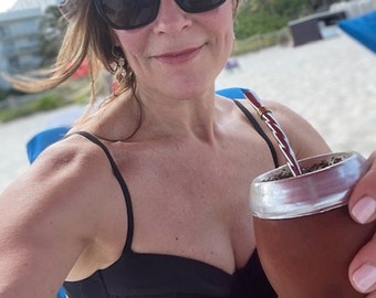 May include: A woman wearing a black bikini top and a floral bucket hat is sitting on a beach chair and holding a silver and brown mate cup with a red and white striped straw.