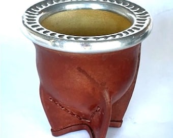 May include: A brown leather cup with a silver rim. The cup has a rounded bottom and three legs.