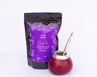 May include: A black pouch of "soul maté" organic yerba maté tea with a purple label and a red gourd with a metal straw. The label reads "Original" with leaf illustrations. The gourd is round and has a silver rim.