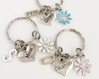 May include: Three silver keychains with charms. The charms include a heart with the word "Mum", a heart with the word "Gran", a heart with the word "Daughter", a silver letter "S", a silver letter "D", a white flower with a rhinestone center, a blue flower with a rhinestone center, and a pink flower with a rhinestone center.