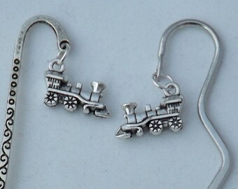 May include: Two silver metal bookmarks. One bookmark has a decorative swirl design and a train charm. The other bookmark has a wavy design and a train charm. The train charms are small and detailed.