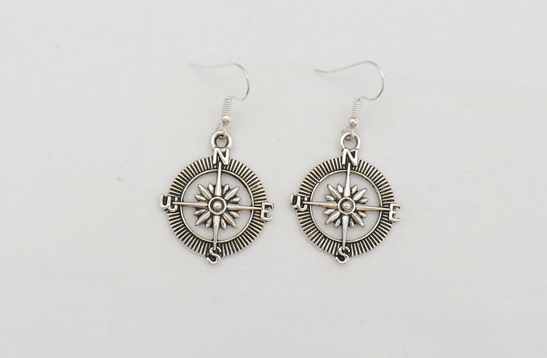 May include: A pair of silver compass earrings with a starburst design and the letters N, S, E, and W around the edge.