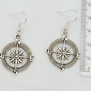 May include: A pair of silver compass earrings with a detailed design. The compass features the cardinal directions N, S, E, and W.