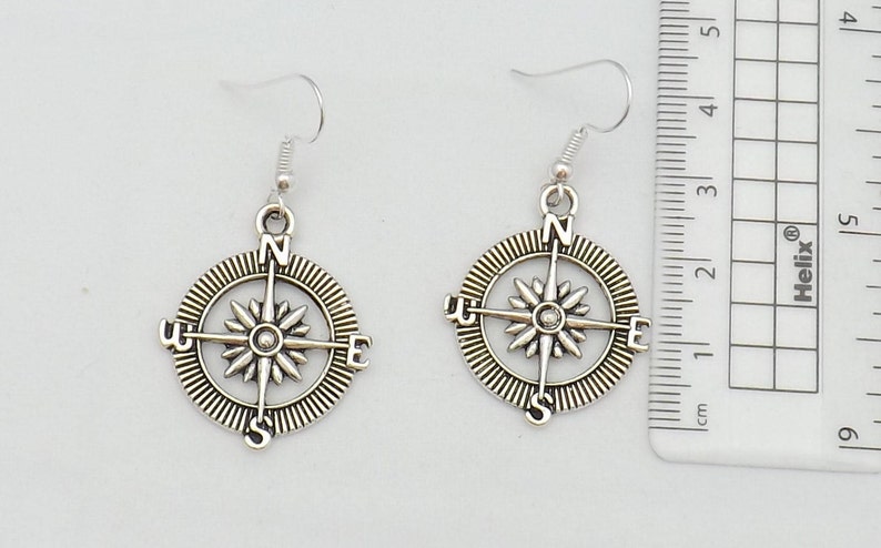 May include: A pair of silver compass earrings with a detailed design. The compass features the cardinal directions N, S, E, and W.