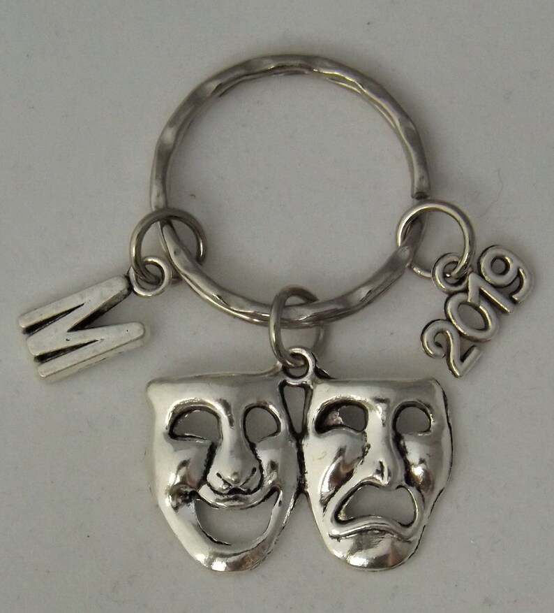 May include: Silver key chain with a silver charm of two theater masks, one smiling and one frowning. The keychain also has a silver charm with the letter "M" and another charm with the year "2019".