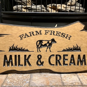 May include: A wooden sign with the text "FARM FRESH MILK & CREAM" carved into it. A black silhouette of a cow is in the center of the sign, with grass carved below it.