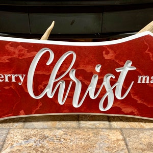 May include: Red wooden sign with white lettering that reads "merry Christ mas".