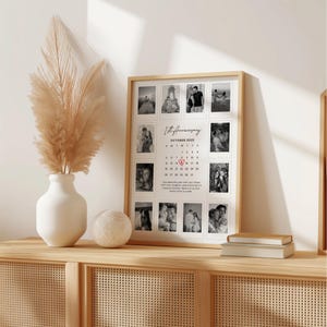 May include: A framed 1st Anniversary photo collage with a light wood frame. The collage features twelve black and white photos arranged around a calendar for October 2025. A vase with pampas grass and a decorative ball sit on a wooden cabinet.