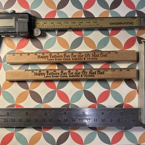 May include: Two wooden rulers with the text "Happy Fathers Day for our DIY Mad Dad Love from Llana, Isabella & Victoria" printed on them. The rulers are on a patterned background with a silver caliper, a metal ruler, and a metal protractor.