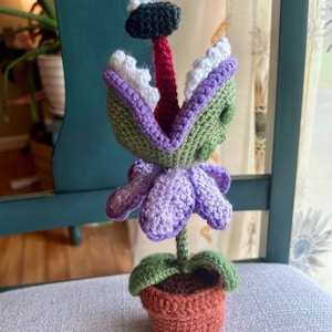 May include: A handmade crochet plant with a large, open mouth, purple and green leaves, and a red tongue. The plant is in a small, orange pot. The plant's mouth is open, revealing white and purple teeth. The plant is sitting on a chair.