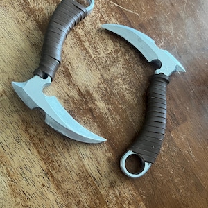 May include: Two silver karambit knives with curved blades and brown wrapped handles. Each knife has a finger ring at the end of the handle. The blades are silver and the handles are wrapped in brown material.