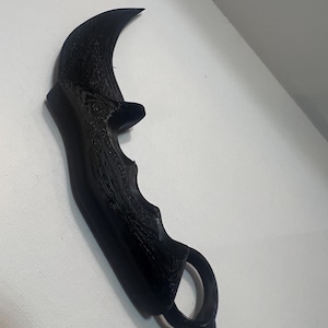 Könnte beinhalten: Ein schwarzes Karambit-Messer mit gebogener Klinge und Fingerring. Der Griff hat eine strukturierte Oberfläche. Das Messer ist gegen eine weiße Wand geneigt.