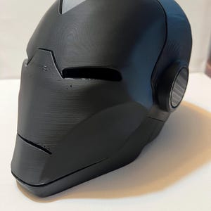 May include: A black, 3D-printed helmet with a smooth, matte finish. The helmet features eye slits, a chin, and a circular detail on the side. The top of the helmet has a grey accent.