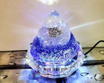 May include: A clear glass crystal with a swirling design, filled with purple glitter and a white light inside. The crystal is on a clear glass base with a light-up feature.