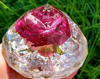 May include: A clear, faceted, diamond-shaped resin paperweight with a preserved pink rose inside. The paperweight is filled with iridescent glitter.
