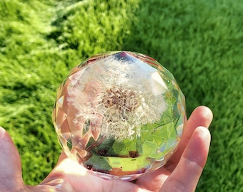 May include: A clear glass sphere with a faceted design, containing a preserved dandelion flower. The sphere is held in a hand against a green grassy background.