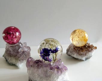 May include: Three clear resin spheres containing dried flowers, each mounted on a rough amethyst crystal cluster. One sphere holds pink flowers, one holds blue and white flowers, and the third contains yellow botanical elements.
