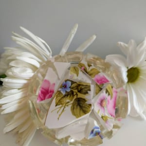 May include: A white ceramic coaster with a floral design. The coaster is made of broken pieces of china that have been glued together. The coaster is surrounded by white flowers.