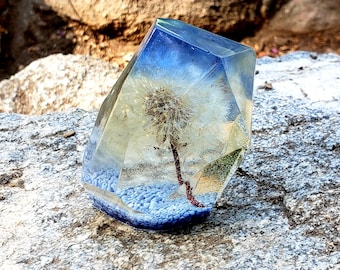 May include: A clear glass prism with a dandelion flower preserved inside. The prism has a blue and white gradient and is sitting on a gray rock.