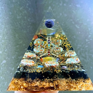 May include: A clear glass pyramid with a gold base and a blue sphere at the top. Inside the pyramid are seven chakra stones, a gold-colored metal figure, and black and gold glitter.
