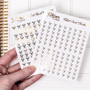 May include: Two sheets of planner stickers with gold foil accents. One sheet features a repeating pattern of bows and the other sheet features a repeating pattern of hearts. Both sheets have the text "I Adore Planning" and "Foiled Heart Checklist" printed on them.