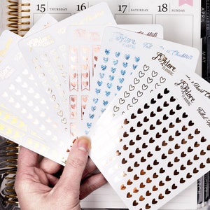 May include: A set of planner stickers with a heart theme. The stickers are in various colors, including gold, rose gold, and blue. The stickers feature different designs, including hearts, banners, and the text "I Adore Planning".