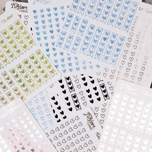 May include: Assortment of planner stickers with heart designs. The stickers are in various colors, including gold, blue, black, white, and pink. The stickers feature different designs, including hearts, banners, and checklists.