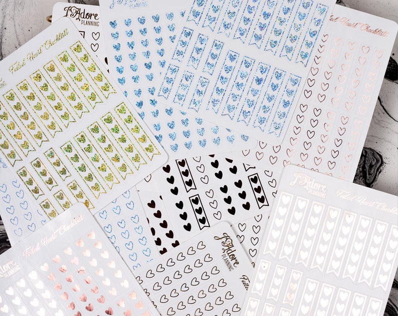 May include: Assortment of planner stickers with heart designs. The stickers are in various colors, including gold, blue, black, white, and pink. The stickers feature different designs, including hearts, banners, and checklists.