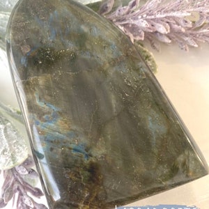 May include: A large, dark gray labradorite crystal with blue and green iridescent flashes. The crystal is shaped like a teardrop and has a smooth, polished surface. The word "LABRADORITE" is written in white letters on a blue background below the crystal.