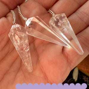 Puede incluir: Tres péndulos de cristal de cuarzo transparente con cadenas plateadas que cuelgan de una mano. CLEAR QUARTZ PENDULUMS está escrito en blanco sobre un fondo morado.