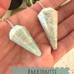 May include: Two green amazonite crystal pendulums with silver chains. The pendulums are in the shape of a point and have a light green color with white streaks. The text "Amazonite Pendulums" is written in white on a green background.