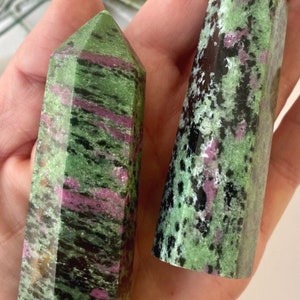 May include: Two polished ruby zoisite obelisks. The stones feature a mix of green, black, and pink hues. The text "RUBY ZOISITE" is visible in a pink font.