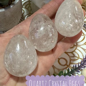 May include: Three clear quartz crystal eggs in the shape of an egg. The eggs are smooth and have a cloudy appearance. The eggs are being held in a hand.