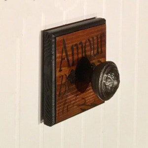 May include: A wooden sign with the word "Amour" written in black paint. The sign is mounted on a wall with a black metal knob.