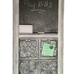 May include: A white framed chalkboard with a black chalkboard surface that says "Pay Bills" in white chalk. The chalkboard has a small white rectangular space below it. Below the chalkboard is a decorative metal tile surface with four tiles. A green sticky note with the text "Dentist appt Monday 2:30 pm" is attached to the metal tile surface.