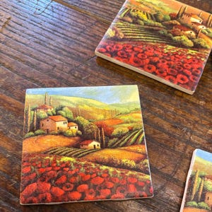 May include: Square ceramic coasters with a Tuscan landscape design. The coasters feature a vibrant scene of rolling hills, vineyards, and red poppy fields. The artwork includes houses and trees under a blue sky. These coasters are perfect for home decor.