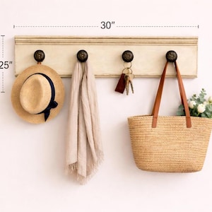 May include: A cream-colored wall-mounted coat rack, 30 inches wide and 9.25 inches high, with four dark bronze knobs. A straw hat with a navy blue band, a beige scarf, keys, and a woven tote bag with flowers hang from the knobs.
