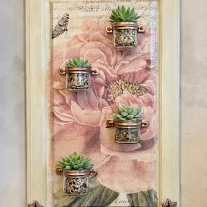 May include: A rectangular wall decor piece with four small succulent plants in glass jars with copper-colored holders. The background features a large pink floral design with script writing and a butterfly. The frame is off-white.