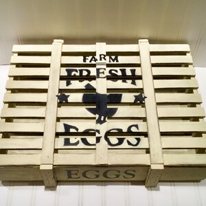May include: A white wooden crate with black lettering that reads "FARM FRESH EGGS" and a black silhouette of a chicken. The crate has a distressed look and is perfect for storing or displaying eggs.